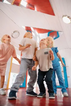 Fire barn står på et rødt tregulv og ser opp i et lyst rom med hvitt tak, lamper og fargerike vinduer.