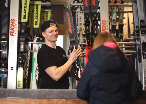 En mann i skiutleien holder et par alpinski, mens en kunde i jakke står ved disken foran ham.
