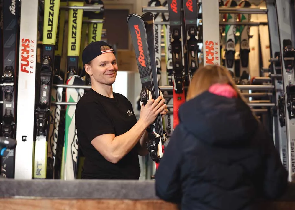 En mann i skiutleien holder et par alpinski, mens en kunde i jakke står ved disken foran ham.